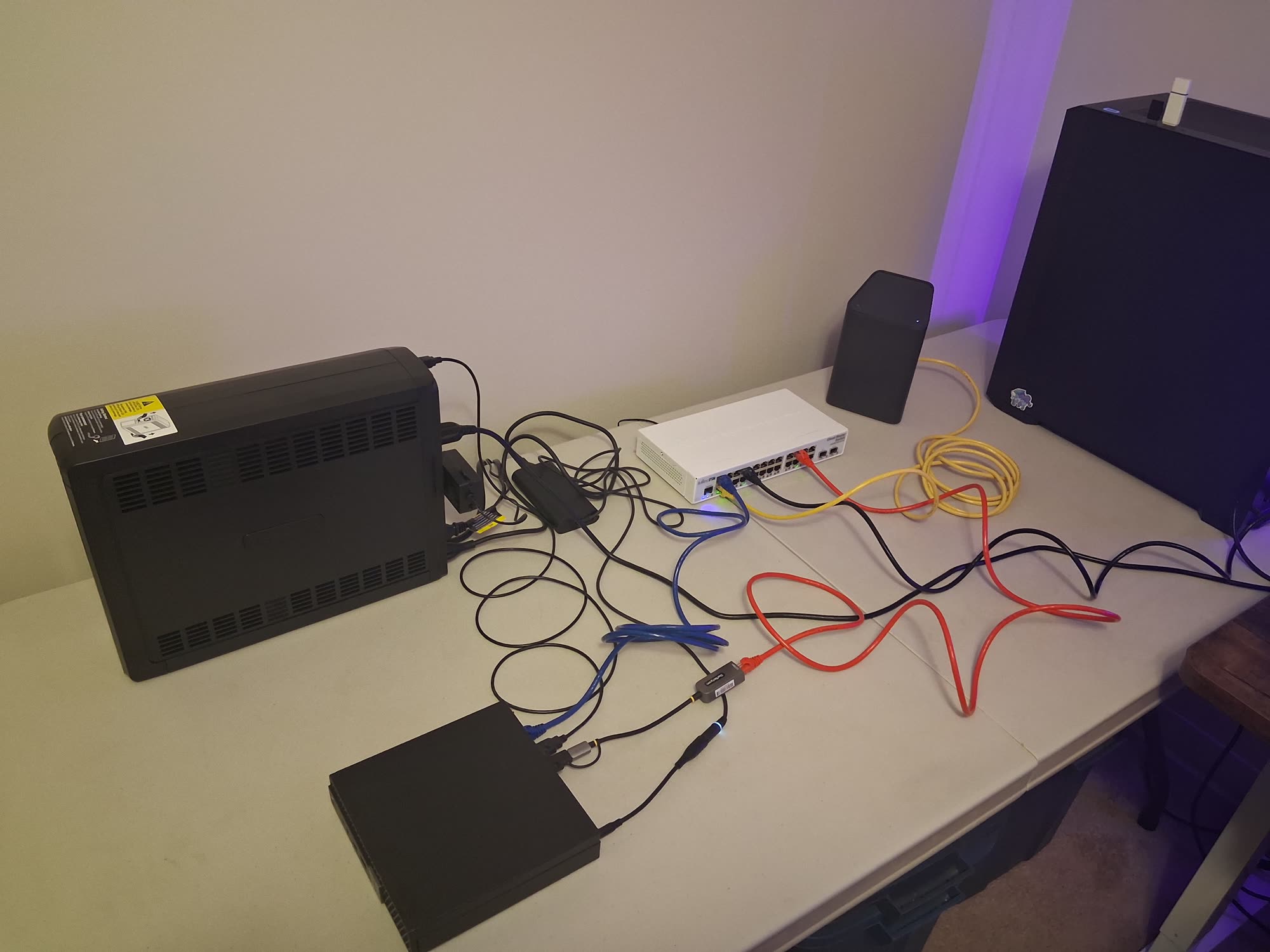 The homelab hardware on a bench: uninterruptible power supply, managed switch, hypervisor, ISP gateway and desktop, wired with colour-coded cables.